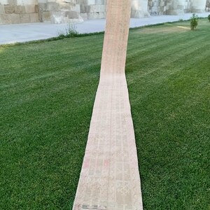Può includere: Un lungo tappeto rettangolare con un motivo sbiadito nei toni del beige, rosa e rosso. Il tappeto è esposto all'aperto su un prato verde, con un edificio in pietra sullo sfondo. Le dimensioni del tappeto non sono visibili.
