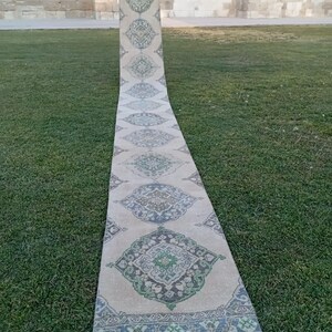 May include: A long, rectangular rug with a repeating geometric pattern in beige, green, and blue. The rug is displayed outdoors on green grass, with a stone wall in the background. The rug's design features diamond-shaped medallions.