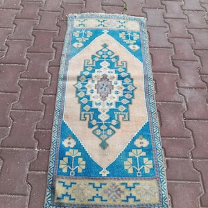 May include: A rectangular woven rug featuring a central diamond medallion in shades of blue, cream, and lavender. The design includes geometric floral motifs and a border with repeating patterns, finished with short fringe at both ends.