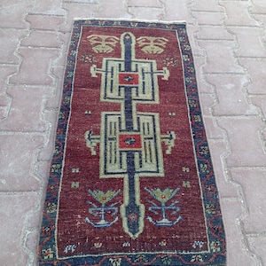 Könnte beinhalten: Ein rechteckiger Vintage-Teppich mit tiefrotem Hintergrund und komplizierten geometrischen Mustern in Blau-, Grün- und Beigetönen. Der Teppich hat ein zentrales Design mit einer Reihe von verbundenen Formen und floralen Motiven an den Enden. Maße: ca. 60 x 90 cm.