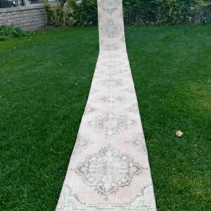 Könnte beinhalten: Ein langer, rechteckiger Teppich mit verblasstem, Vintage-Aussehen. Der Teppich weist ein zentrales Design mit floralen und geometrischen Mustern in Rosa-, Beige- und Olivgrüntönen auf. Der Teppich wird im Freien auf einem grünen Rasen präsentiert.