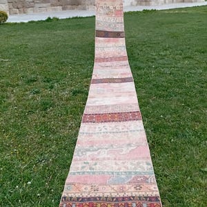 Peut inclure: Un long tapis étroit et vintage avec un motif patchwork. Le tapis présente une variété de couleurs, notamment du rose, du rouge, du marron et du beige. Le tapis est exposé à l'extérieur sur de l'herbe verte.