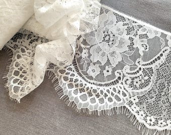 May include: White lace trim with a floral design and scalloped edges. The lace is rolled up, with a portion of the trim spread out, showcasing the intricate details and texture. The trim is on a gray surface.