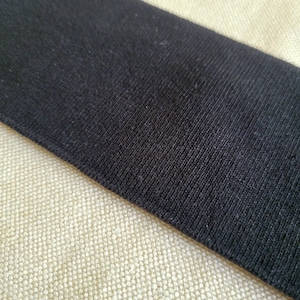 May include: Close-up of a black fabric strip against a beige fabric background. The black fabric has a textured, woven appearance.
