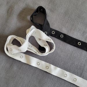 May include: Two rolls of white cotton webbing with metal grommets. One roll is laid out flat, the other is rolled up. A black cotton webbing with metal grommets is laid out flat.