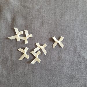 Pode incluir: Uma coleção de pequenas fitas de cetim de cor marfim espalhadas sobre uma superfície de tecido cinzento. As fitas são amarradas em um estilo clássico, com laços e caudas limpas. O tecido tem uma textura sutil, proporcionando um fundo neutro para as fitas delicadas.