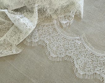 May include: A length of off-white Chantilly lace trim featuring a delicate floral pattern with scalloped edges and fine eyelash fringe detailing. The intricate, sheer fabric displays a complex network of woven threads and openwork motifs.