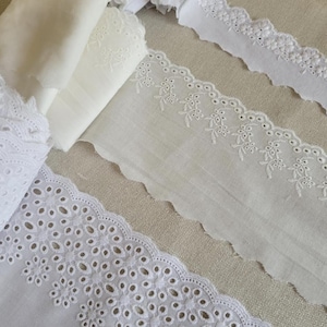 May include: Three white cotton lace trims with scalloped edges and different embroidery patterns. The lace trims are arranged on a beige fabric background.