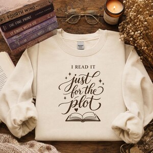 Op de afbeelding: Een crèmekleurig sweatshirt met ronde hals en donkerbruine tekst met de tekst I READ IT just for the plot, vergezeld van een klein hartpictogram en een grafische illustratie van een open boek met decoratieve sterren rondom de tekst.