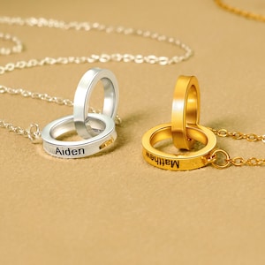 May include: A silver and gold necklace with two interlocking rings. The rings are engraved with the names "Aiden" and "Matthew".