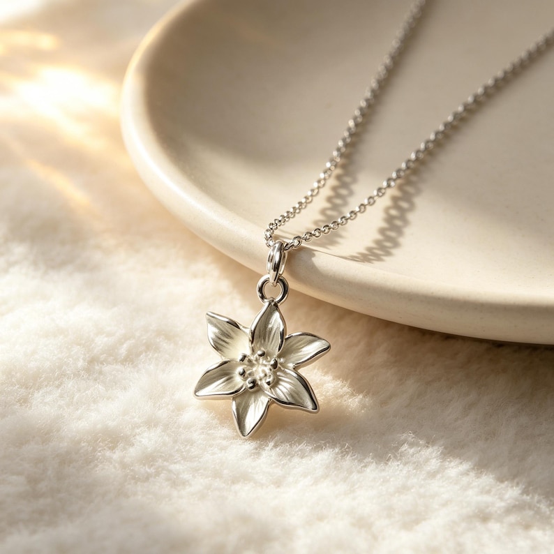 May include: A silver-colored flower pendant necklace. The pendant is a detailed, six-petal flower with a small, round center. The necklace chain is delicate and hangs on a white, textured surface.