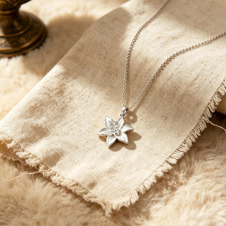 May include: A silver-colored necklace with a flower pendant. The pendant is a detailed, six-petal flower with a small, textured center. The necklace chain is delicate and hangs on a beige fabric, resting on a fluffy, light-colored surface.