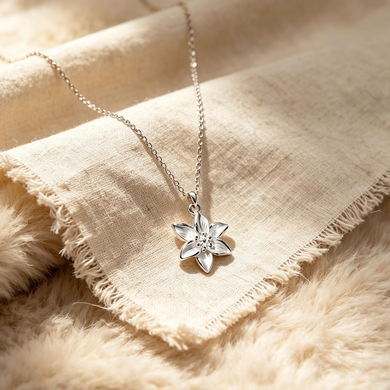 May include: A silver-colored necklace with a flower pendant. The pendant has six petals and small, round embellishments in the center. The necklace rests on a beige fabric and a fluffy, light brown surface.