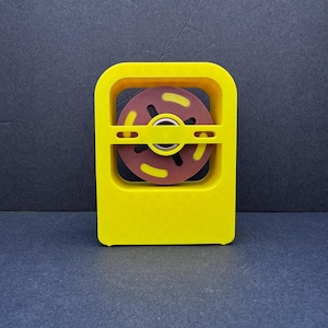 May include: A yellow plastic device with a brown and yellow spinning disc inside. The disc has a central hole and a series of spokes.