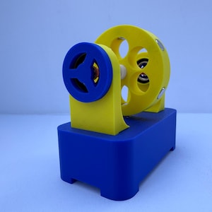 May include: A 3D-printed machine in primary colors. The base is blue, supporting a yellow structure with a circular wheel featuring multiple holes. A blue wheel is on the side. The design suggests a mechanical or educational toy.