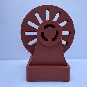 May include: A red plastic spinning wheel with a base. The wheel has 12 slots and a center hub. The base is rectangular with rounded corners.