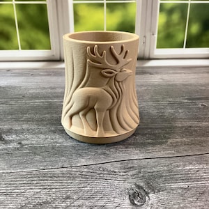 May include: A light brown wooden pencil holder with a carved design of a deer. The cylindrical holder has a detailed relief of a deer and tree bark. The pencil holder is approximately 4 inches tall and 3 inches in diameter.