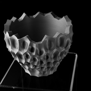 May include: A grayscale ceramic bowl with a textured, honeycomb-like exterior and a jagged rim. The bowl is displayed on a clear, angled stand against a black background. The bowl's design features a series of rounded, hexagonal shapes.