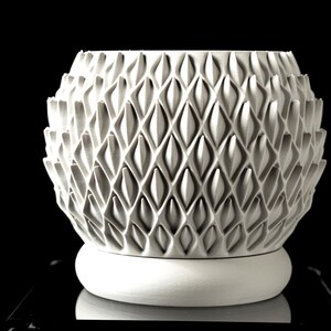 May include: A white, textured decorative vase with a geometric pattern. The vase has a bulbous shape with a series of diamond-shaped indentations. It sits on a round, white base. The background is black.
