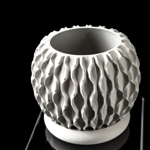 May include: A white, textured ceramic vase with a spherical shape and a wide opening. The vase features a repeating wave-like pattern around its exterior and sits on a circular base. The background is black.