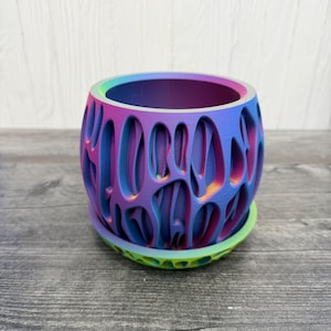 May include: A vibrant, multi-colored plant pot with a unique, openwork design. The pot transitions from purple to blue and green, with a matching saucer. The design features an intricate pattern of oval openings, creating a modern, artistic look.