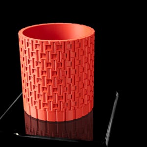 May include: A cylindrical, coral-colored container with a repeating geometric pattern. The container has a textured surface and is set against a black background. The design features a series of rectangular and square shapes.