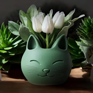 May include: A green ceramic cat planter with a smiling face and whiskers. The planter is filled with white tulips.