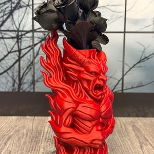 May include: A red, devil-themed vase with a detailed, muscular design. The vase holds a bouquet of black fabric roses. The vase has a fiery, sculpted appearance, with the devil's face and horns prominently displayed.