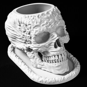 May include: A white, textured skull-shaped planter with a wide opening at the top and a tray-like base. The skull features detailed facial features, including eye sockets, a nose cavity, and teeth. The planter is set against a black background.