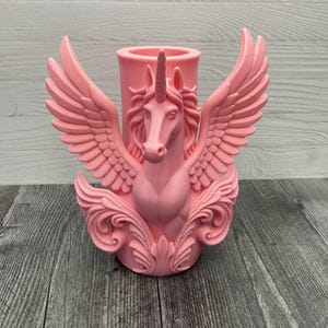 May include: A pink unicorn-themed candle holder. The candle holder features a unicorn head with a horn, wings, and decorative flourishes. The wings are spread wide, and the candle holder is a solid pink color. The background is a wooden surface.