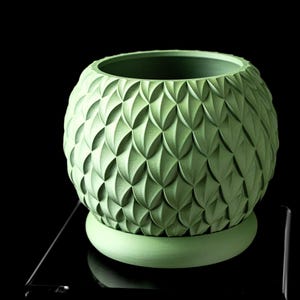 May include: A green, spherical planter with a textured, repeating pattern. The planter sits on a matching round base. The design features a series of overlapping, leaf-like shapes. The planter is made of a solid, matte material.