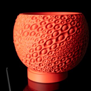 May include: A vibrant orange decorative bowl with a textured, organic pattern. The bowl sits on a matching circular base, creating a striking contrast against the black background. The design features a series of raised, oval-shaped details across its surface.