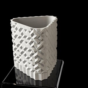 May include: A white, geometric vase with a textured surface. The vase has a rectangular shape with a triangular opening and is displayed on a black surface. The design features repeating geometric patterns.