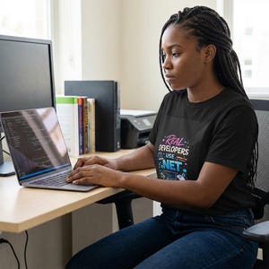 May include: A black t-shirt with the text "Real Developers Use .NET" in a colourful design. The person is sitting at a desk, working on a laptop computer. A computer monitor, books, and a printer are also visible.
