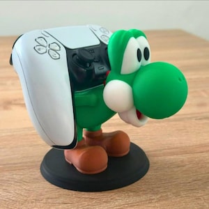 May include: A green Yoshi-themed controller stand. The stand features a white PlayStation 5 controller held by a green Yoshi figure. The figure has a large green snout, white eyes, and brown shoes. The stand is on a black circular base.