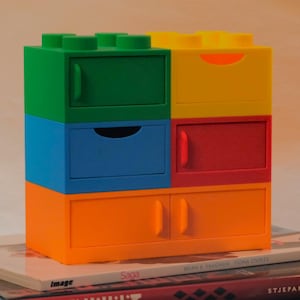Puede incluir: Un mueble de almacenamiento colorido que se asemeja a una pila de bloques de construcción de juguete. La unidad presenta cajones en verde, azul, amarillo, rojo y naranja. La unidad está colocada encima de una pila de libros.