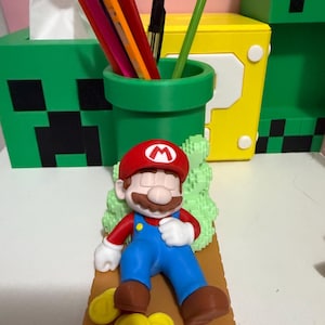 May include: A desk organizer set featuring a green pipe pencil holder, a green Minecraft-themed tissue box, and a yellow question mark box. A Mario figurine rests on a brown base with yellow coins. Pencils and a pen are in the pencil holder.