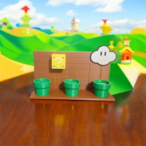 May include: A miniature diorama featuring elements from a video game. It includes three green pipes, a brown brick wall with a question mark block, and a white cloud. The background depicts a stylized landscape with hills and buildings.