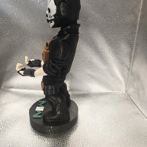 K&ouml;nnte beinhalten: Eine schwarze Figur eines Charakters mit Totenkopfmaske, der eine schwarze Jacke und Stiefel tr&auml;gt. Die Figur steht auf einem schwarzen Sockel mit den Buchstaben "MW" in Gr&uuml;n. Die Figur hat einen beigefarbenen Werkzeugg&uuml;rtel und Handschuhe.