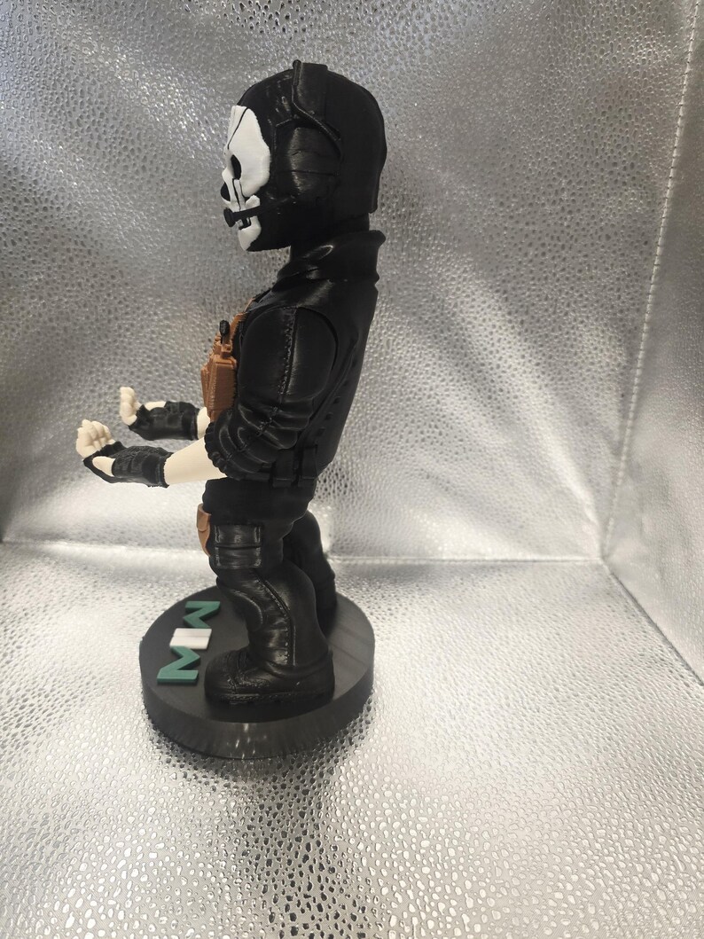 K&ouml;nnte beinhalten: Eine schwarze Figur eines Charakters mit Totenkopfmaske, der eine schwarze Jacke und Stiefel tr&auml;gt. Die Figur steht auf einem schwarzen Sockel mit den Buchstaben "MW" in Gr&uuml;n. Die Figur hat einen beigefarbenen Werkzeugg&uuml;rtel und Handschuhe.