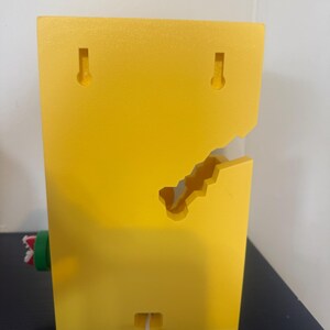 Pode incluir: Um objeto retangular amarelo brilhante montado na parede com um design recortado exclusivo. Possui duas ranhuras em forma de fechadura para pendurar e uma pequena abertura na parte inferior com um cabo branco saindo. Uma pe&ccedil;a cil&iacute;ndrica verde e vermelha est&aacute; presa na lateral.