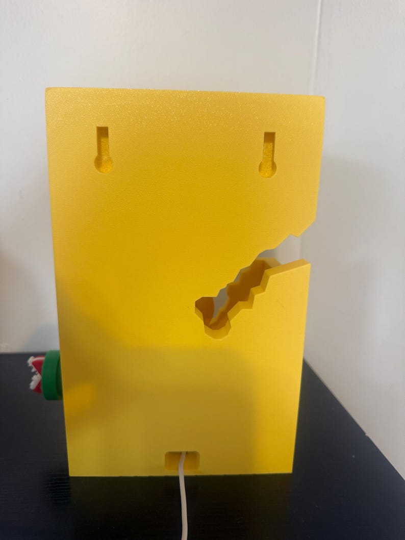 Pode incluir: Um objeto retangular amarelo brilhante montado na parede com um design recortado exclusivo. Possui duas ranhuras em forma de fechadura para pendurar e uma pequena abertura na parte inferior com um cabo branco saindo. Uma pe&ccedil;a cil&iacute;ndrica verde e vermelha est&aacute; presa na lateral.