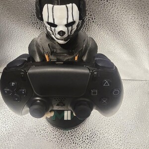 K&ouml;nnte beinhalten: Eine schwarz-wei&szlig;e Figur eines Charakters mit Headset und Totenkopfmaske, der einen schwarzen Gamecontroller h&auml;lt. Der Controller hat Kn&ouml;pfe und Joysticks. Die Figur steht auf einem schwarzen Sockel mit den Buchstaben "M" und "II".
