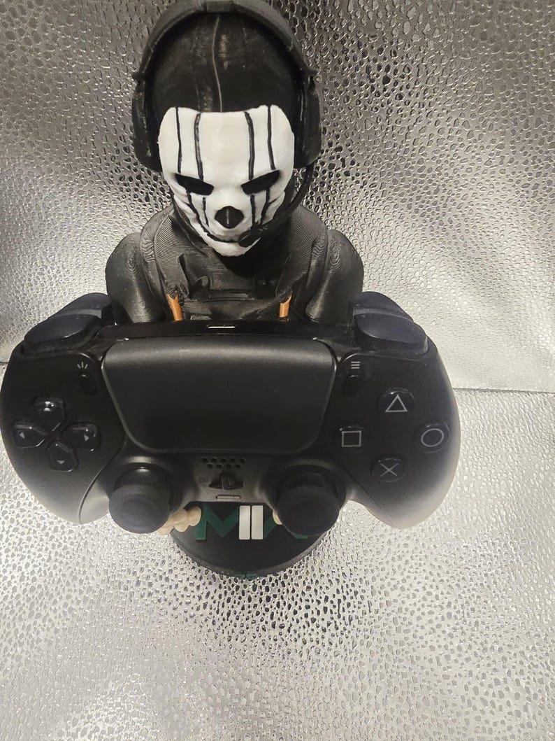 K&ouml;nnte beinhalten: Eine schwarz-wei&szlig;e Figur eines Charakters mit Headset und Totenkopfmaske, der einen schwarzen Gamecontroller h&auml;lt. Der Controller hat Kn&ouml;pfe und Joysticks. Die Figur steht auf einem schwarzen Sockel mit den Buchstaben "M" und "II".