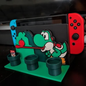 May include: A green and white Yoshi-themed Nintendo Switch game cartridge holder. The holder features a green base with three green pipes and a black background with a Yoshi character design. A red and blue Switch controller is visible.