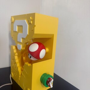 Pode incluir: L&acirc;mpada retangular amarela com design de ponto de interroga&ccedil;&atilde;o e cogumelo vermelho e branco. A l&acirc;mpada tem uma planta Piranha verde e vermelha na lateral. Um cabo branco se estende da base.