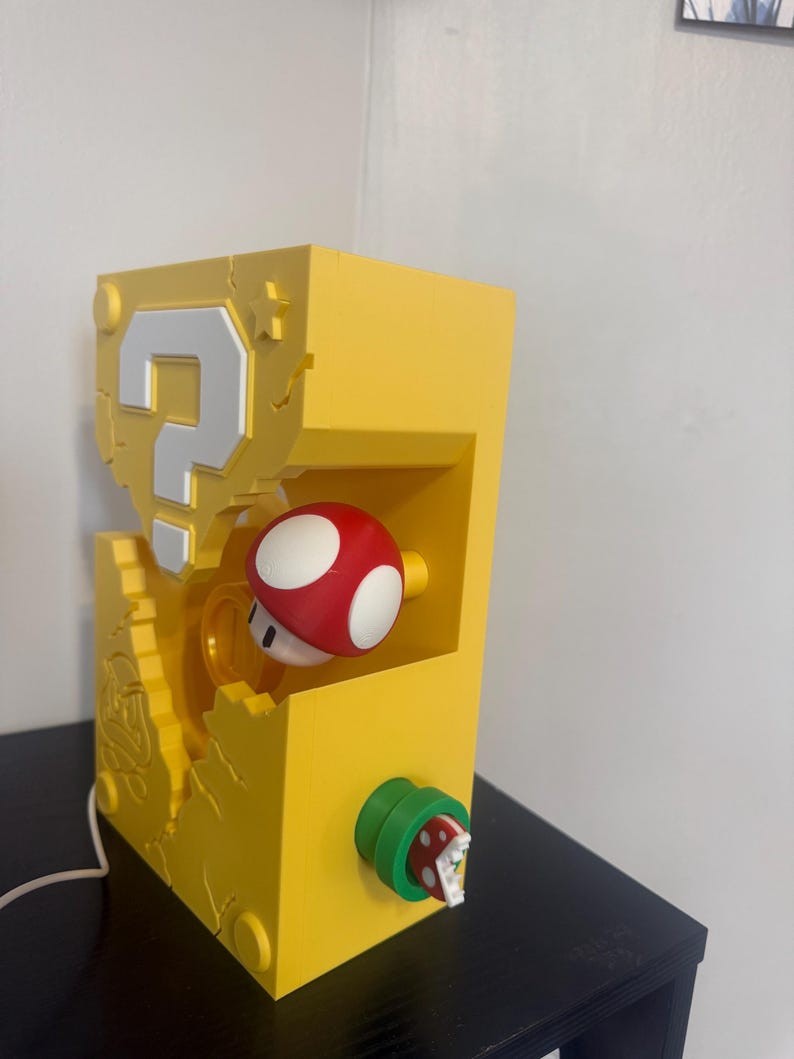 Pode incluir: L&acirc;mpada retangular amarela com design de ponto de interroga&ccedil;&atilde;o e cogumelo vermelho e branco. A l&acirc;mpada tem uma planta Piranha verde e vermelha na lateral. Um cabo branco se estende da base.