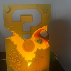 Pode incluir: L&acirc;mpada amarela iluminada em forma de caixa de ponto de interroga&ccedil;&atilde;o, com design de moeda, cogumelo e Goomba. A l&acirc;mpada tem uma apar&ecirc;ncia rachada e uma estrela na lateral. &Eacute; um item decorativo.