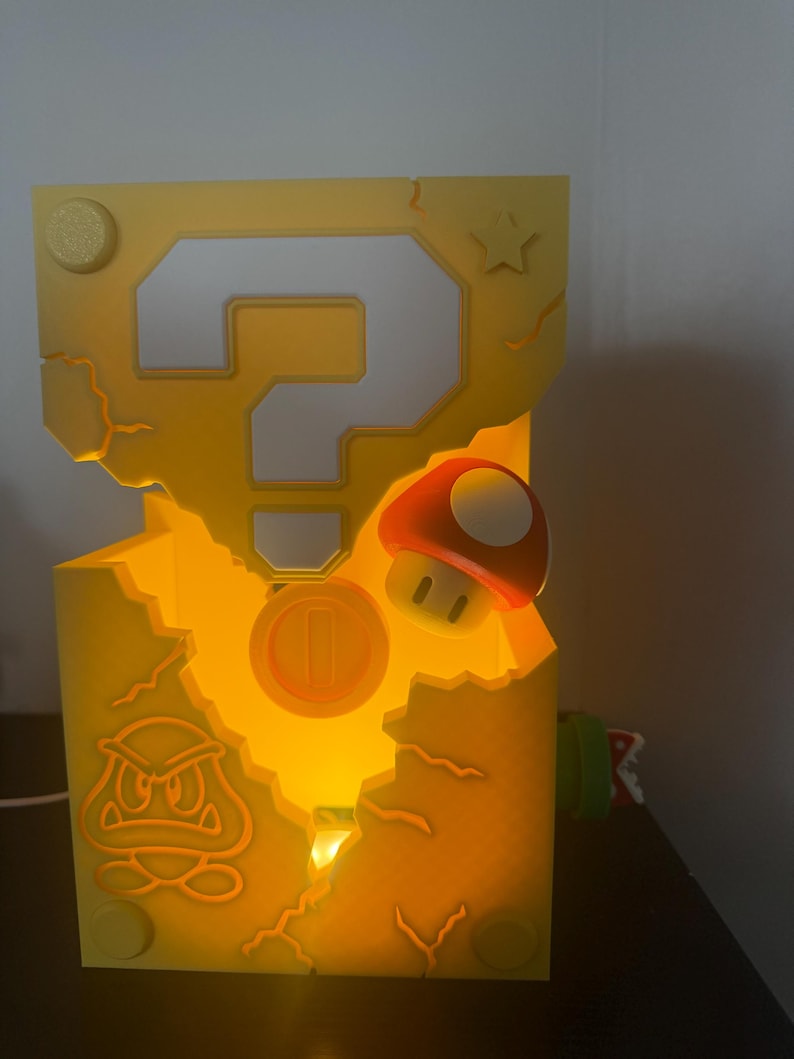 Pode incluir: L&acirc;mpada amarela iluminada em forma de caixa de ponto de interroga&ccedil;&atilde;o, com design de moeda, cogumelo e Goomba. A l&acirc;mpada tem uma apar&ecirc;ncia rachada e uma estrela na lateral. &Eacute; um item decorativo.