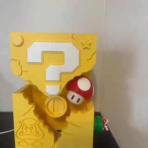 Pode incluir: Um bloco amarelo com um ponto de interroga&ccedil;&atilde;o branco, um cogumelo vermelho e branco e uma moeda de ouro. O bloco tem um design Goomba e uma estrela. O objeto &eacute; impresso em 3D.
